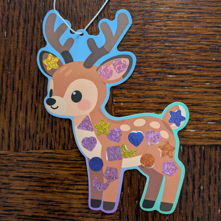 Cerf en carton décoré de gommettes pailletées violettes, dorées et bleues, posé sur une table en bois avec sa ficelle de suspension.