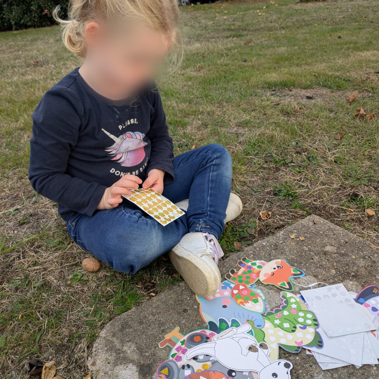 Petite fille assise dans l'herbe, une planche de gommettes dorées à la main, entourée d'animaux en carton déjà décorés.